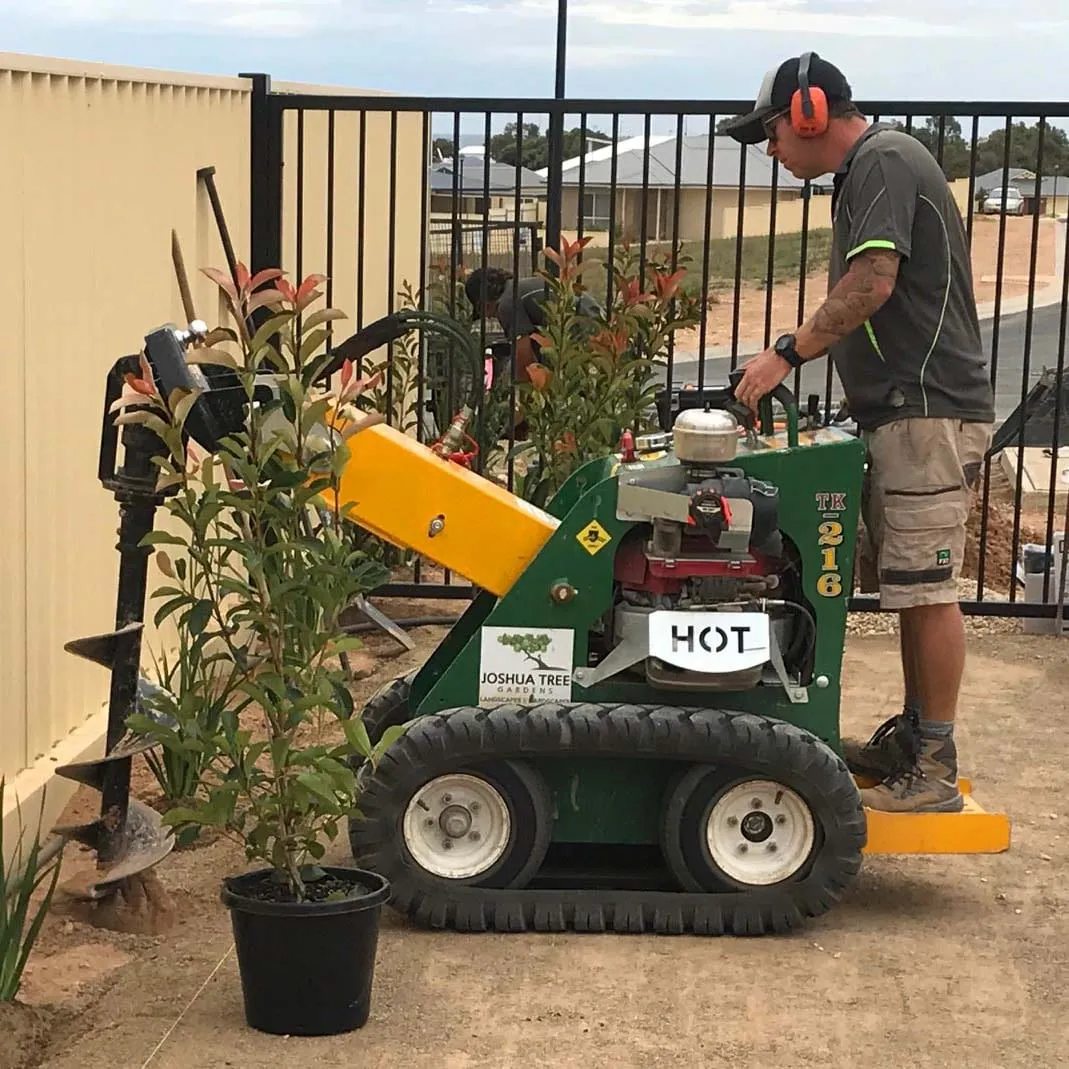A Joshua Tree Landscaping mini loader working in a backyard beside a fence, with an auger attachment on the front