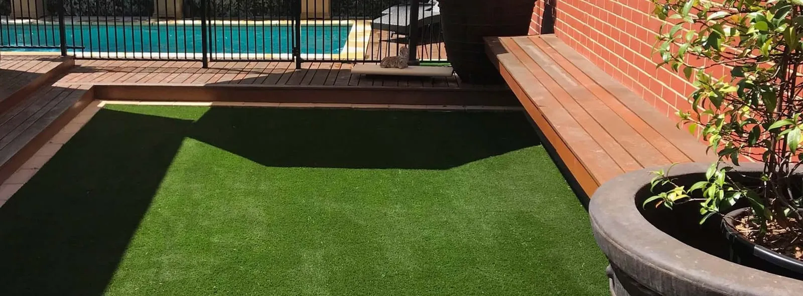 A finished backyard with artificial turf laid flush to timber decking beside a pool, with a brick planter along one side