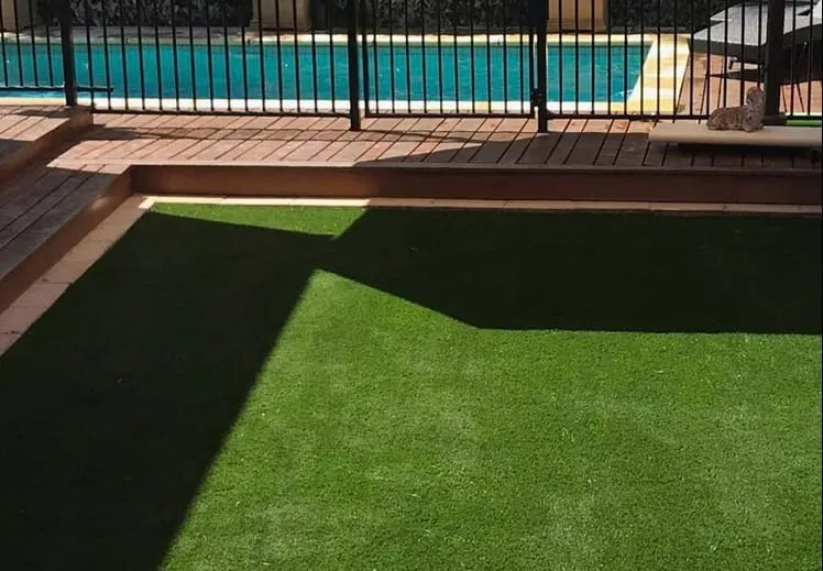 Artificial turf laid flush against timber decking beside a swimming pool, with a brick planter along one edge