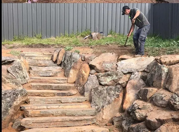 Stone steps running up beside a rock retaining wall, with a worker raking the bed alongside